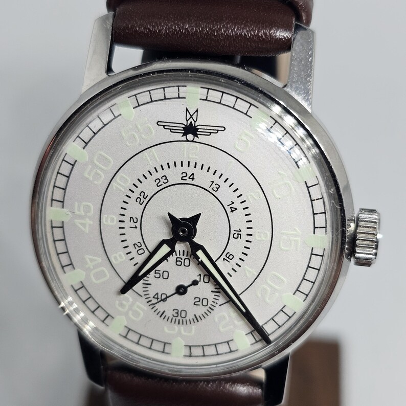 K&ouml;nnte beinhalten: Eine silberfarbene Armbanduhr mit einem braunen Lederarmband. Das Zifferblatt der Uhr ist wei&szlig; mit schwarzen Ziffern und Zeigern sowie einem kleinen Sekundenzeiger. Die Uhr hat eine Krone zum Aufziehen und Einstellen der Zeit. Das Armband ist ca. 20 cm lang.