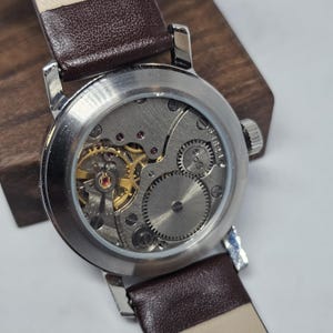 K&ouml;nnte beinhalten: Nahaufnahme einer Vintage-Armbanduhr mit silbernem Geh&auml;use und braunem Lederarmband. Der Uhrenr&uuml;cken zeigt das komplizierte mechanische Uhrwerk mit goldfarbenen Zahnr&auml;dern und silbernen Komponenten. Das Armband ist etwa 2 cm breit.