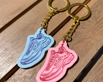 Puede incluir: Dos llaveros con dijes en forma de zapatillas deportivas unidos a anillas de metal dorado. Un dije es azul claro con una ilustración de zapatilla azul oscuro, y el otro es rosa claro con una ilustración de zapatilla rosa oscuro. Ambos incluyen el texto RUN WITH IT.