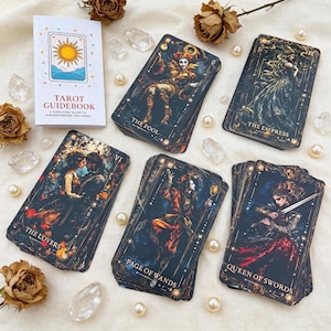 以下が含まれることがあります： The Fool、The Empress、The Lovers、Page of Wands、Queen of Swordsとラベル付けされた5つのカードの山と、白いTarot Guidebook。カードには、金色のアクセントと白い真珠をあしらった、暗く複雑なイラストが描かれています。