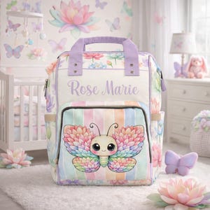 Puede incluir: Un bolso de pañales blanco y pastel con un diseño de mariposa. El bolso presenta una gran ilustración de mariposa en la parte delantera, con un fondo a rayas. El nombre "Rose Marie" está escrito en la parte delantera del bolso. El bolso tiene un asa y detalles morados.