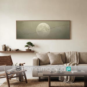 May include: A rectangular framed print featuring a full moon and a line of birds against a hazy backdrop. The artwork hangs above a beige sofa in a living room. A wooden coffee table and chair are in the foreground, creating a neutral-toned space.