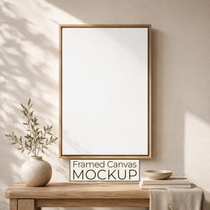 May include: A vertical rectangular blank white canvas inside a thin light brown wooden frame. The frame is mounted on a wall. Below the frame, a light-coloured ceramic vase with green leaves sits on a wooden table next to a small bowl and books.