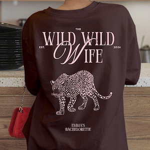 Puede incluir: Una sudadera marr&oacute;n oscuro con el texto "THE WILD WILD WIFE" y una ilustraci&oacute;n de leopardo rosa. El texto "EST. 2026" tambi&eacute;n est&aacute; impreso en la sudadera. El texto "EMMA'S BACHELORETTE" est&aacute; impreso debajo.
