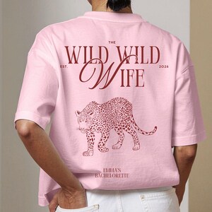 Puede incluir: Camiseta rosa claro con el texto "THE WILD WILD WIFE" y una ilustraci&oacute;n de leopardo. El texto "EST. 2026" est&aacute; encima de "WIFE". Debajo del leopardo est&aacute; el texto "EMMA'S BACHELORETTE".