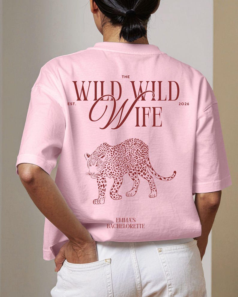 Puede incluir: Camiseta rosa claro con el texto "THE WILD WILD WIFE" y una ilustraci&oacute;n de leopardo. El texto "EST. 2026" est&aacute; encima de "WIFE". Debajo del leopardo est&aacute; el texto "EMMA'S BACHELORETTE".