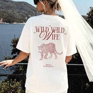 Puede incluir: Camiseta blanca con un dise&ntilde;o de leopardo burdeos y el texto "The Wild Wife" y "Emma's Bachelorette". La camiseta es de corte holgado.