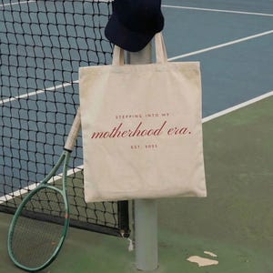 May include: A canvas tote bag with the text "Stepping into my motherhood era. Est. 2023" in red script. A navy blue cap hangs above the bag. A green tennis racket and yellow tennis balls are also visible.