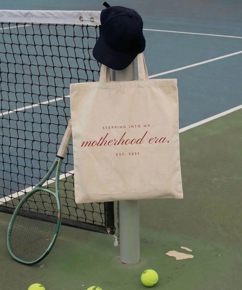 May include: A canvas tote bag with the text "Stepping into my motherhood era. Est. 2023" in red script. A navy blue cap hangs above the bag. A green tennis racket and yellow tennis balls are also visible.