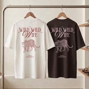 Puede incluir: Dos camisetas, una blanca y otra marr&oacute;n oscuro, colgadas de una barra. Cada camiseta presenta el texto "THE WILD WILD WIFE" y una ilustraci&oacute;n de leopardo en color burdeos. El texto "EMMA'S BACHELORETTE" tambi&eacute;n est&aacute; impreso en las camisetas.
