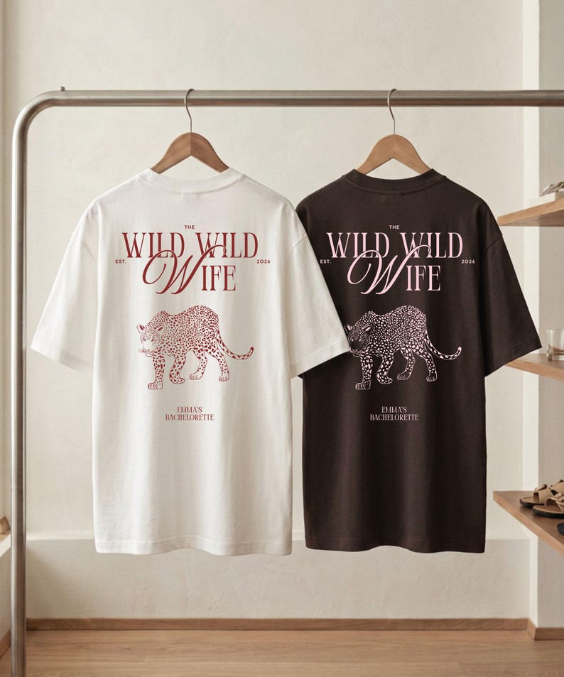 Puede incluir: Dos camisetas, una blanca y otra marr&oacute;n oscuro, colgadas de una barra. Cada camiseta presenta el texto "THE WILD WILD WIFE" y una ilustraci&oacute;n de leopardo en color burdeos. El texto "EMMA'S BACHELORETTE" tambi&eacute;n est&aacute; impreso en las camisetas.