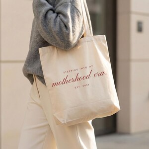 May include: A beige canvas tote bag with the text "STEPPING INTO MY motherhood era. EST. 2025" in burgundy script. The bag is carried over the shoulder. The person is wearing a gray sweater and cream-colored pants.