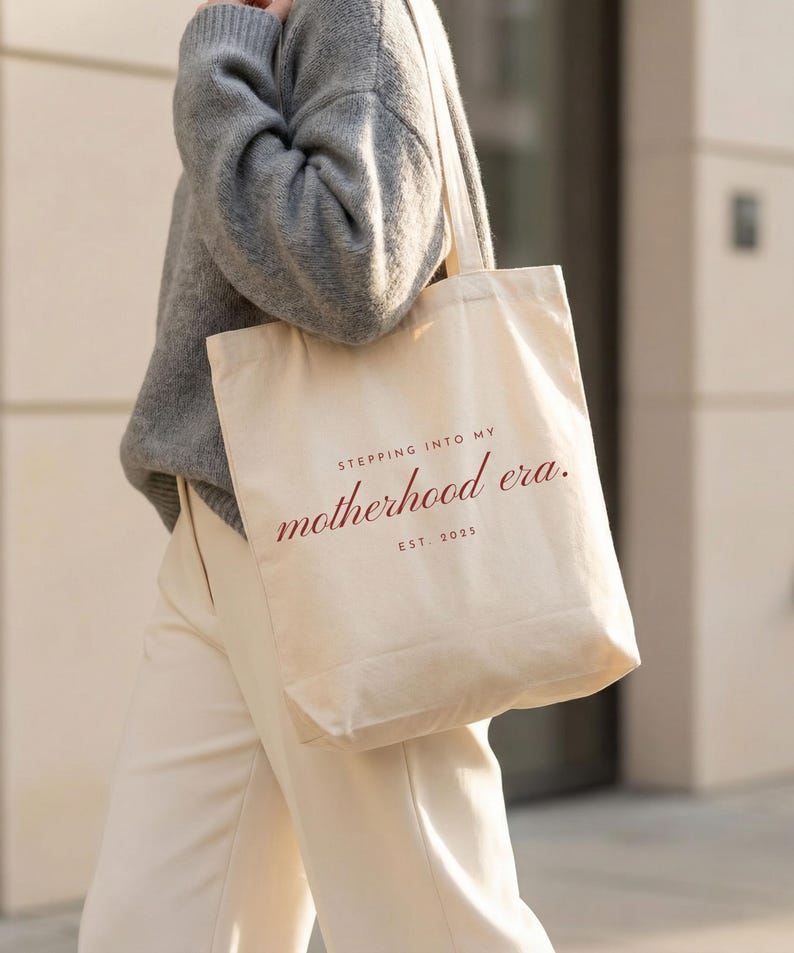 May include: A beige canvas tote bag with the text "STEPPING INTO MY motherhood era. EST. 2025" in burgundy script. The bag is carried over the shoulder. The person is wearing a gray sweater and cream-colored pants.