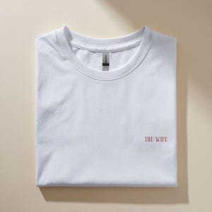 Puede incluir: Una camiseta blanca doblada con el texto "THE WIFE" en rojo en la parte delantera. La camiseta est&aacute; hecha de un material suave y est&aacute; cuidadosamente doblada. El fondo es de color beige neutro.