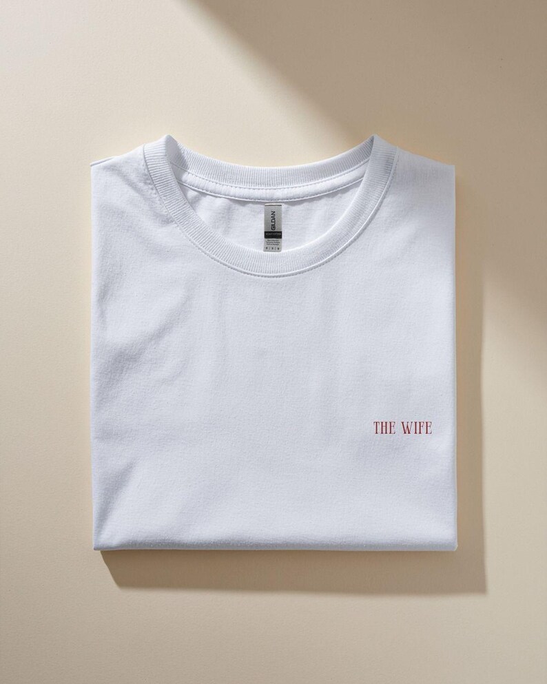 Puede incluir: Una camiseta blanca doblada con el texto "THE WIFE" en rojo en la parte delantera. La camiseta est&aacute; hecha de un material suave y est&aacute; cuidadosamente doblada. El fondo es de color beige neutro.
