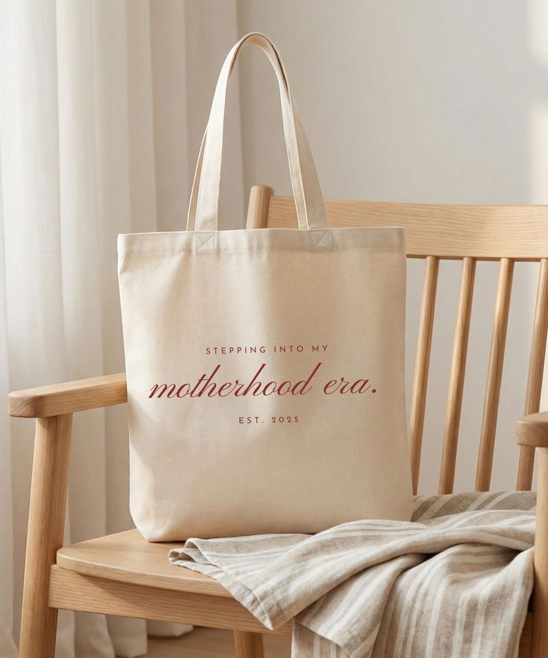 May include: A cream-colored tote bag with the text "STEPPING INTO MY motherhood era. EST. 2025" in a burgundy script. The bag is sitting on a wooden chair with a striped cloth draped over it.