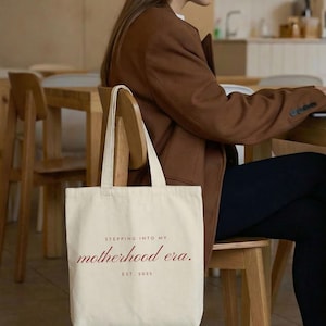 May include: Cream-colored tote bag with the text "Stepping into my motherhood era. Est. 2025" in a burgundy script. The bag is held by a person wearing a brown blazer and black pants, seated at a wooden table.