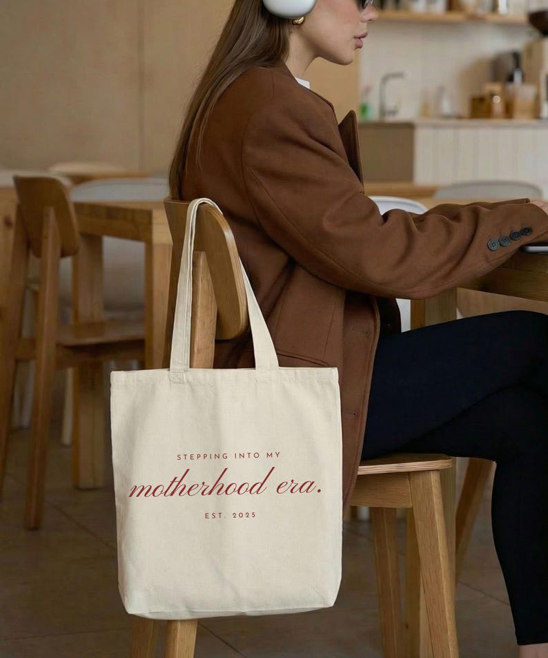 May include: Cream-colored tote bag with the text "Stepping into my motherhood era. Est. 2025" in a burgundy script. The bag is held by a person wearing a brown blazer and black pants, seated at a wooden table.