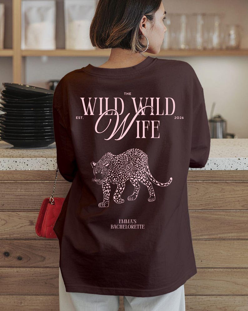 Puede incluir: Una sudadera marr&oacute;n oscuro con el texto "THE WILD WILD WIFE" y una ilustraci&oacute;n de leopardo rosa. El texto "EST. 2026" tambi&eacute;n est&aacute; impreso en la sudadera. El texto "EMMA'S BACHELORETTE" est&aacute; impreso debajo.