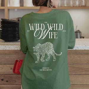 Puede incluir: Camiseta verde de manga larga con texto blanco y un dise&ntilde;o de leopardo. El texto dice "THE WILD WILD WIFE" con "EST. 2026" y "EMMA'S BACHELORETTE" debajo de la ilustraci&oacute;n del leopardo. La camiseta es de color verde liso.
