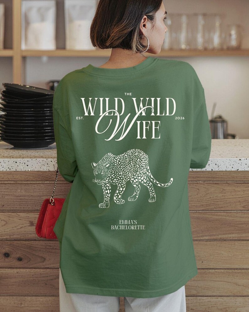 Puede incluir: Camiseta verde de manga larga con texto blanco y un dise&ntilde;o de leopardo. El texto dice "THE WILD WILD WIFE" con "EST. 2026" y "EMMA'S BACHELORETTE" debajo de la ilustraci&oacute;n del leopardo. La camiseta es de color verde liso.