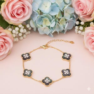 May include: A gold-colored bracelet featuring five clover-shaped charms with a black and white design. The bracelet is set against a soft pink background with pink roses and blue hydrangeas.