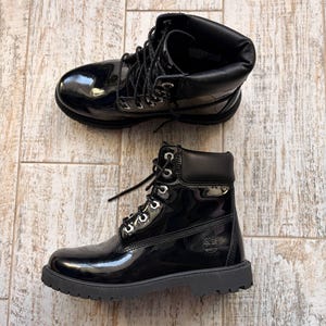 Puede incluir: Dos botas negras de charol con cordones. Las botas tienen un acabado brillante y una suela gruesa negra. El logotipo de Timberland es visible en el lateral de una bota. Las botas se exhiben sobre una superficie clara con vetas de madera.