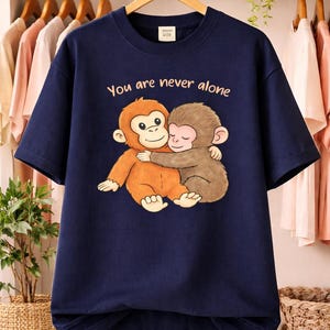 May include: A navy blue t-shirt with a cartoon illustration of two monkeys embracing. The text "You are never alone" is printed above the monkeys. The t-shirt is displayed on a wooden hanger.