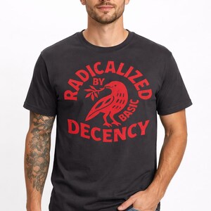以下が含まれることがあります： 赤色のグラフィックが入った濃いグレーのTシャツ。グラフィックは枝を持った鳥で、「RADICALIZED BY BASIC DECENCY」という言葉に囲まれています。このシャツはクルーネックで、青いジーンズと組み合わせています。
