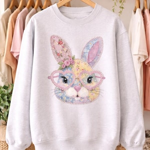 May include: A light gray sweatshirt featuring a colorful bunny face with heart-shaped glasses. The bunny has floral accents on its ears and head. The design is a patchwork of pastel colors, perfect for Easter or spring.