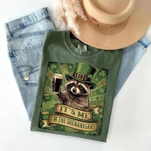 Puede incluir: Camiseta verde con un mapache con un sombrero verde de duende y una pinta de cerveza. La camiseta tiene el texto "IT'S ME I'M THE SHENANIGANS" en una pancarta. La imagen incluye vaqueros azules y un sombrero beige.