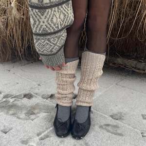 May include: A person wearing beige knitted leg warmers and black ballet flats. The leg warmers are ribbed and have a textured pattern. A gray and white patterned sweater is visible in the image.