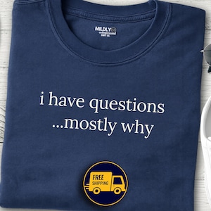 Puede incluir: Camiseta azul marino con el texto blanco "i have questions...mostly why". La camiseta tiene cuello redondo y un pequeño logo. También se ven un sombrero de paja, gafas de sol y zapatillas blancas.