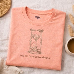 Puede incluir: Camiseta color melocotón con un gráfico de un reloj de arena lleno de calaveras. El texto "i do not have the bandwidth" está impreso debajo. La camiseta tiene cuello redondo y la marca "Mildly Unimpressed Shirt Co." está impresa arriba a la derecha.
