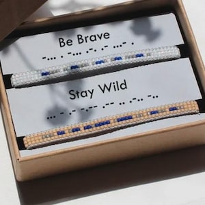 Puede incluir: Una caja de regalo contiene dos pulseras de cuentas con las frases "Be Brave" y "Stay Wild" en código Morse. Las pulseras son blancas y beige con cuentas azules y plateadas. La caja es marrón claro.