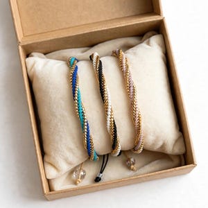 Peut inclure: Trois bracelets torsadés composés de perles dorées métalliques entrelacées avec des perles mates bleu et turquoise, noir et blanc, et rose pâle. Chaque bracelet possède une petite perle transparente au bout du cordon réglable, présenté en boîte.