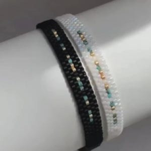 Puede incluir: Dos pulseras de cuentas. Una es negra con detalles turquesa y dorados, la otra es blanca con detalles turquesa y dorados. Las pulseras están una al lado de la otra, mostrando el intrincado trabajo de cuentas y el diseño.