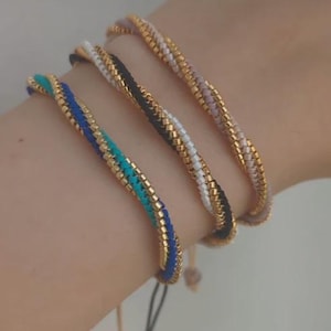 Puede incluir: Cuatro pulseras de cuentas en tonos azul, turquesa, negro, blanco y rosa. Las pulseras tienen un borde de metal dorado y se muestran en una muñeca. Cada pulsera tiene una combinación de colores diferente y un cierre de cordón.