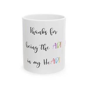 May include: A white ceramic mug featuring the black text Thanks for being the ART in my HeART. The letters ART are highlighted in various colours including purple, blue, green, yellow, and red, creating a vibrant contrast against the white surface.
