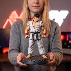 Könnte beinhalten: Eine detaillierte Figur des Street Fighter-Charakters Ryu, dargestellt mit kurzem schwarzem Haar, einem roten Stirnband und einem weißen Karate-Gi mit schwarzem Gürtel. Die Figur steht auf einem dunklen, strukturierten runden Sockel.