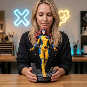 May include: A collectible figurine of a female superhero with long red hair, wearing a yellow and blue bodysuit with a circular emblem on the chest and belt. The figure stands on a dark gray, textured base with a red X symbol on the front.