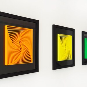 Puede incluir: Tres obras de arte enmarcadas con un diseño geométrico en espiral. El arte presenta colores vibrantes: naranja, amarillo y verde. Cada pieza está encerrada en un marco negro, creando una exhibición moderna. La obra de arte está montada en una pared blanca.