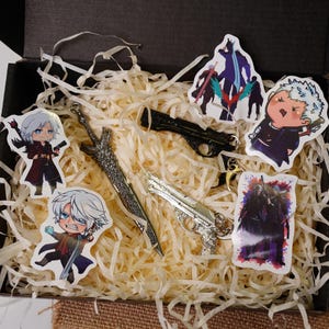 Könnte beinhalten: Eine Geschenkbox gefüllt mit Dekopapier, mit Charakteraufklebern und Metall-Schlüsselanhängern. Die Schlüsselanhänger umfassen zwei Pistolen-Designs und ein Schwert. Die Aufkleber zeigen Anime-Charaktere in verschiedenen Posen.