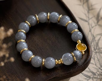 May include: A beaded bracelet featuring smooth, round, translucent grey beads alternating with small, polished gold-toned metal spacers. One prominent, circular, translucent amber-colored bead is set within a gold-toned metal frame.