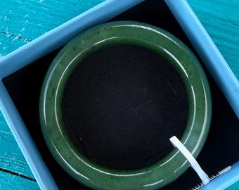May include: A smooth, circular dark green bangle bracelet with subtle natural inclusions, presented inside a square light blue box with a black velvet interior lining. A small white plastic tag is attached to the side of the bangle.
