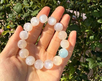 May include: A stretch bracelet composed of smooth, round, translucent white and pale blue moonstone beads. The beads exhibit a subtle iridescent sheen and are strung together in a circular formation, creating a continuous loop of polished stones.