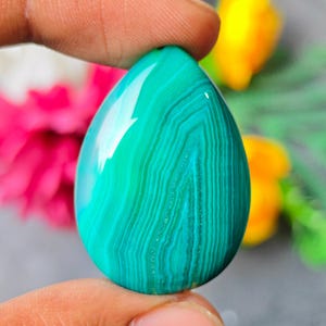 Op de afbeelding: Een traanvormige edelsteen met levendige blauwgroene en groene banden met een glad, gepolijst oppervlak. De steen vertoont ingewikkelde, natuurlijke concentrische patronen door de hele structuur en reflecteert het licht met een glanzende afwerking.