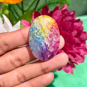Op de afbeelding: Een ovaalvormige cabochon edelsteen met een gebarsten textuur en een levendig regenboogverloop van gele, blauwe, paarse en roze tinten. De steen heeft een glad, gepolijst oppervlak dat de ingewikkelde, webachtige patronen goed laat uitkomen.