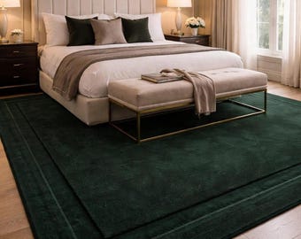 May include: A large, rectangular, deep forest green area rug with a subtle border detail, positioned on a light-colored wooden floor beneath a bed frame, a tufted bench with a gold metal base, and bedroom furniture in a neutral-toned room.
