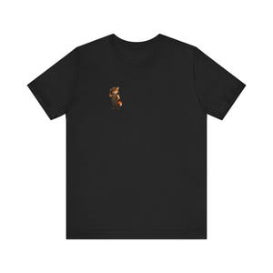 Pode incluir: Uma t-shirt preta de manga curta com um pequeno gráfico centrado na parte superior esquerda do peito. O gráfico retrata uma personagem raposa antropomórfica a usar um chapéu castanho e roupa escura, numa pose casual com a mão levantada.
